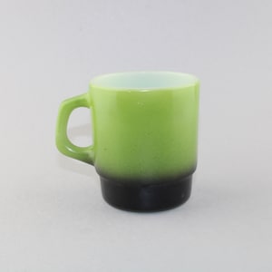 Puede incluir: Una taza de estilo vintage con un degradado de color verde y negro. La taza tiene un asa redondeada y una forma cilíndrica. La mitad superior es de un verde brillante, que pasa a negro en la base. La taza es probablemente de cerámica o vidrio.