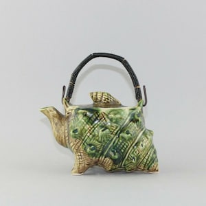 May include: A green and brown ceramic teapot shaped like a seashell with a black wire handle.