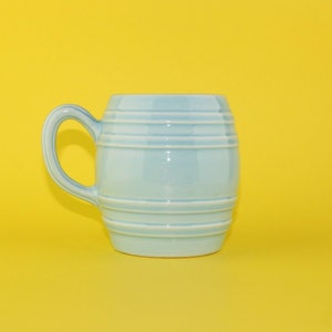 May include: A light blue ceramic mug shaped like a barrel with horizontal lines. The mug has a handle and is sitting on a yellow surface.