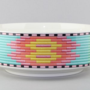 May include: A white ceramic bowl with a colorful geometric pattern around the rim. The pattern features pink, yellow, green, and black stripes.