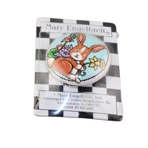 Può includere: Pomello rotondo in ceramica con un coniglio dei cartoni animati che tiene un fiore, su sfondo azzurro. Confezionato su una carta a scacchi bianchi e neri con il testo "Mary Engelbreit".