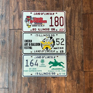 May include: Three vintage license plates from Lincoln, Illinois. The top plate is red and white and says "Land of Lincoln" and "Lincoln Art & Balloon Festival" with the dates August 25, 26, 27 and the number 180. The middle plate is white and says "Land of Lincoln" and "Lincoln Art & Balloon Festival" with the dates August 27-28-29 and the number 152. The bottom plate is white and says "Land of Lincoln" and "Mt. Zion Pony Express Days" with the dates August 13-16, 1998 and the number 164.