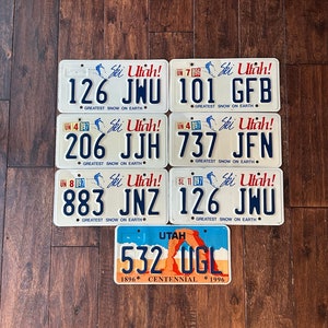 May include: Six vintage Utah license plates with the slogan "Ski Utah! Greatest Snow on Earth."  The plates feature different numbers and letters, including "126 JNU", "101 GFB", "206 JJH", "737 JFN", "883 JNZ", and "126 JWU".  The plates are blue, white, and red.  There is also a Utah license plate with the number "532 UGL" and the text "1896 Centennial 1996".