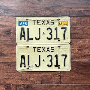 May include: Two vintage Texas license plates with the number ALJ 317. One plate has an "APR" sticker and the other has an "88 Texas" sticker. The plates are cream-colored with black lettering and numbers.
