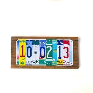 Puede incluir: Una placa de madera con la fecha 10-02-13 escrita con placas de matr&iacute;cula de colores. La placa est&aacute; montada en una pared blanca.