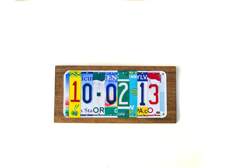 Puede incluir: Una placa de madera con la fecha 10-02-13 escrita con placas de matr&iacute;cula de colores. La placa est&aacute; montada en una pared blanca.