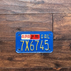 May include: A vintage California license plate with a blue background and yellow lettering. The plate reads "7X6745" and includes a sticker indicating "APR 95". The plate is rectangular with rounded corners.