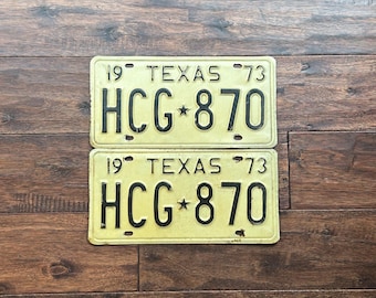 May include: Two vintage yellow Texas license plates with black lettering. The plates read "19 TEXAS 73 HCG*870".
