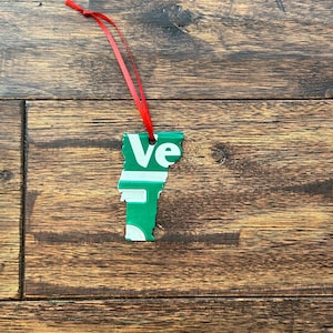 May include: Green and white Vermont state-shaped ornament with a red ribbon. The ornament has the letters "Vt" on it.