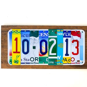 Puede incluir: Una placa de madera con la fecha 10-02-13 escrita con placas de matr&iacute;cula de colores. La placa es de madera y tiene un aspecto r&uacute;stico.
