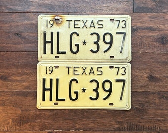 May include: Two vintage Texas license plates from 1973. The plates are yellow with black lettering and numbers, including "HLG 397" and "TEXAS". The plates show signs of wear and tear, with some rust and discoloration.