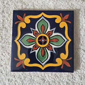 May include: A square ceramic tile with a colorful floral design. The tile features a central flower with red, yellow, and green petals, surrounded by a blue background and a yellow border.