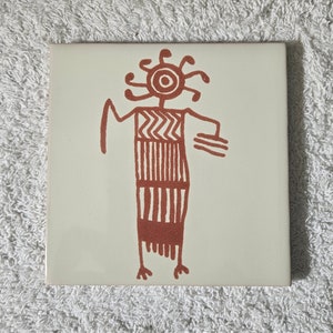 May include: A white ceramic tile with a red-brown stylized drawing of a person with a sun-like head and a patterned dress.