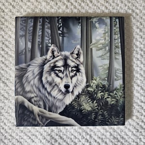 May include: A ceramic tile with a detailed image of a grey wolf in a forest setting. The wolf is looking directly at the viewer with a serious expression. The background is a dark green and brown forest with tall trees and ferns.