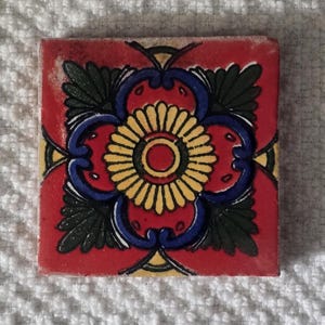 May include: A red, blue, yellow, and green ceramic tile with a floral design. The tile has a central flower with a yellow center and blue petals. The flower is surrounded by green leaves and blue swirls.