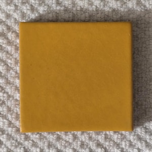 May include: A square, mustard-yellow tile with a slightly textured surface. The tile is set against a white, textured background.