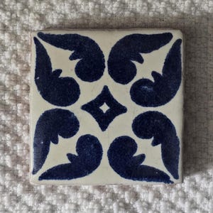 May include: A white ceramic tile with a blue and white geometric pattern. The pattern features four stylized leaves in each corner and a diamond shape in the center.