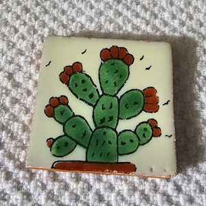 May include: A hand-painted ceramic tile featuring a green cactus with red flowers. The tile has a white background and a brown border.