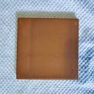 May include: A square terracotta tile with a slightly rough texture. The tile is a warm brown color and has a slightly faded appearance.
