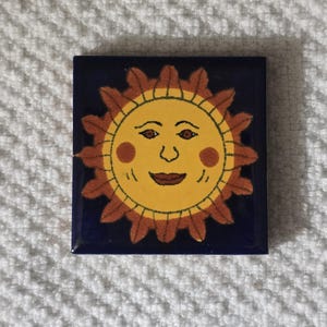 May include: Square ceramic tile with a smiling sun design. The sun has a yellow face with brown eyes, rosy cheeks, and a smiling mouth. The sun's rays are brown, and the tile has a dark blue background.