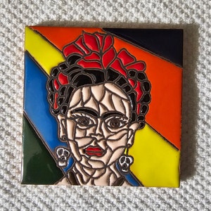 May include: A colorful ceramic tile featuring a portrait of Frida Kahlo. The tile is decorated with a vibrant red, yellow, blue, and green background. Frida Kahlo's face is painted in a white and black mosaic style with a red flower crown.