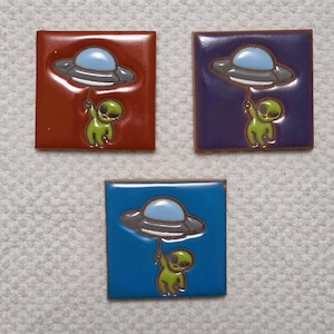 May include: Three square ceramic tiles with a glossy finish. Each tile features a cartoon alien holding a string attached to a flying saucer. The tiles are in red, purple, and blue, with the alien and saucer in complementary colors.