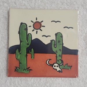 May include: A ceramic tile with a desert scene featuring two green cacti, a red sun, blue mountains, and a white skull with horns. The background is a light beige color with a red sand area.