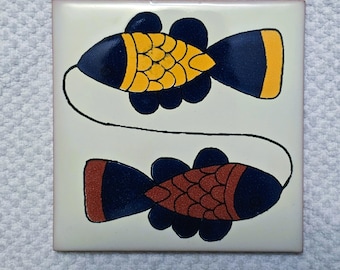 May include: Square ceramic tile featuring two stylized fish. The fish have navy blue bodies with yellow and orange accents. A thin black line connects the two fish. The tile has a cream-colored background.