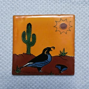 May include: Square ceramic tile with a desert scene. The tile features a black and blue bird, a green cactus, and a yellow sun against an orange sky. The ground is brown, and there is a smaller bird in the scene.