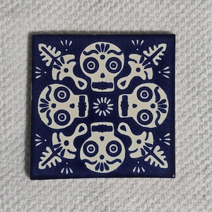 May include: Square ceramic tile with a dark blue background and a repeating pattern of off-white sugar skull designs. The tile features four skulls, floral accents, and a central starburst.