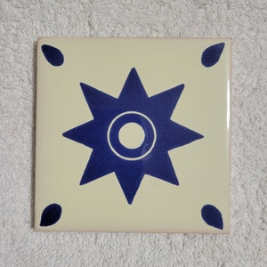 May include: A cream-colored ceramic tile with a blue starburst design. The starburst has a white circle in the center.