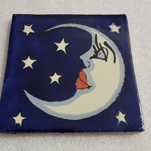 May include: Square ceramic tile with a dark blue background featuring a crescent moon with a face and several white stars. The moon has a light blue outline, a red mouth, and a white face.