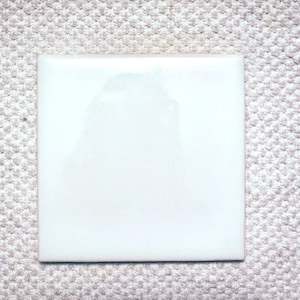 May include: A square, white ceramic tile with a glossy finish. The tile is set against a textured, off-white background. The tile's smooth surface reflects light, creating highlights and shadows.