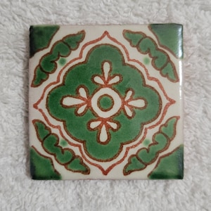 May include: A square ceramic tile with a green and white floral design. The tile has a white background with a green floral pattern in the center. The floral pattern is surrounded by a red border.