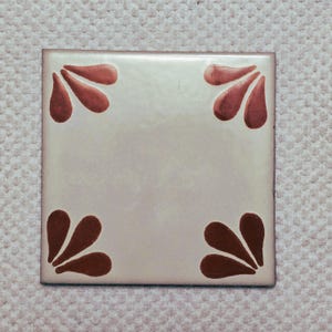 May include: Square ceramic tile with a cream-colored background and four dark red floral corner designs. The tile has a glossy finish and a slightly aged appearance, suitable for home decor.