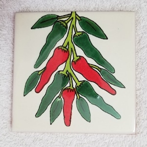 May include: Square ceramic tile with a hand-painted design of chili peppers. The peppers are red and green, with green leaves and stems. The background is off-white. The tile is likely for kitchen or home decor.