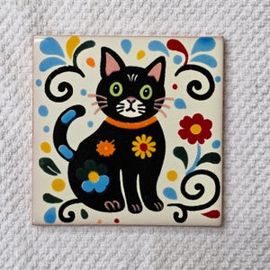 May include: A square ceramic tile featuring a black cat with green eyes and an orange collar. The cat is decorated with orange and yellow flower motifs. The background is off-white and includes colorful floral patterns, blue teardrop shapes, and black swirling lines.