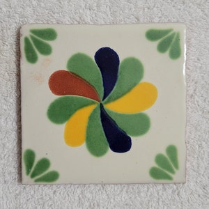 May include: A white ceramic tile with a green, yellow, blue, and orange floral design. The tile has a white background and a green border.