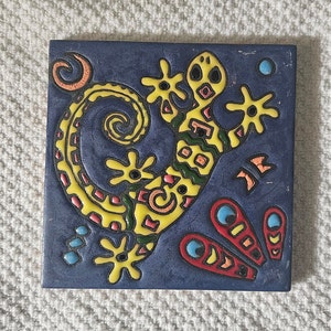 May include: A blue ceramic tile with a yellow, green, and red stylized lizard design. The lizard is outlined in black and has a white background.