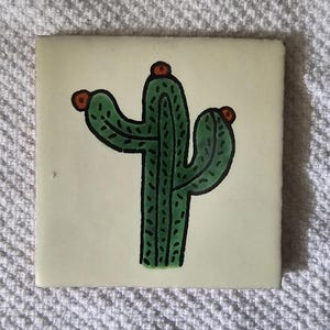 May include: A green cactus with red flowers painted on a white ceramic tile.