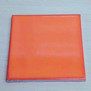 May include: A square, orange ceramic tile with a glossy finish. The tile has a smooth surface and a white edge. The tile is a solid color and appears to be new.