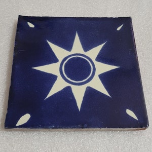 May include: A blue ceramic tile with a white sun design. The sun has eight points and a circle in the center.
