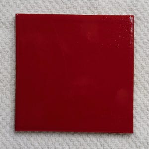 May include: A square tile with a glossy, solid red finish. The surface is smooth and reflective, showing a uniform deep red color across the entire flat face of the object. The edges are straight and defined, forming a perfect four-sided shape.