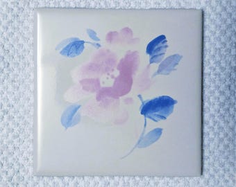 May include: Square ceramic tile with a floral design. The design features a cluster of light purple flowers and blue leaves on a white background. The tile has a glossy finish.