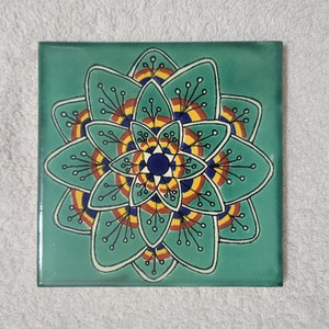 May include: A teal ceramic tile with a colorful mandala design. The mandala features a central circle with concentric rings of yellow, red, and blue, surrounded by a stylized flower with black outlines.