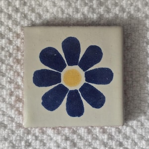 May include: A white ceramic tile with a blue and yellow flower design. The flower has eight petals and a yellow center.