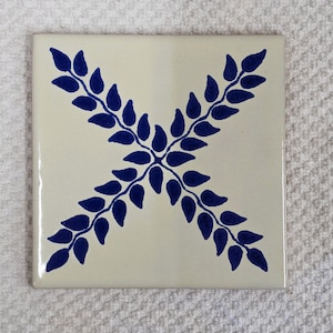 May include: A square ceramic tile with a glossy off-white finish featuring a centered dark blue hand-painted design. The pattern consists of two intersecting leafy branches forming an X shape, with each branch composed of small, pointed leaves.