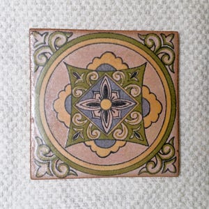 May include: Square ceramic tile with a detailed floral design. The tile features a central diamond shape with a flower, surrounded by a circle and decorative corner elements. Colors include pink, green, yellow, and blue.