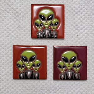 May include: Three square tiles with a glossy finish, each featuring a group of three green alien heads with black eyes and a black suit. The tiles have a red, maroon, and dark red background.