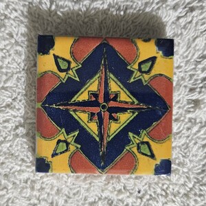 May include: A square ceramic tile with a colorful geometric design. The tile features a central star shape with four points, surrounded by a blue and yellow pattern. The tile is decorated with red, yellow, blue, and green colors.